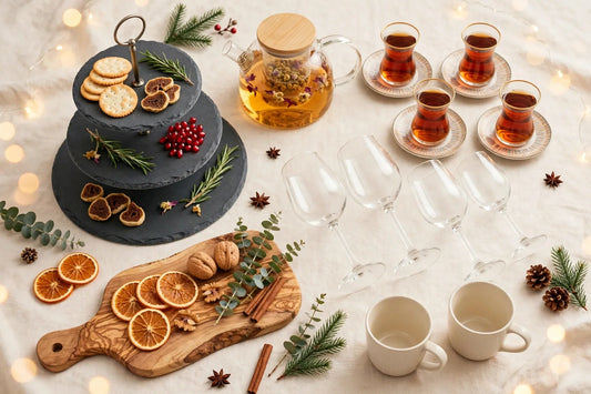 Olive wood charcuterie board, slate tiered serving platter, Turkish tea glasses with saucers, wine glasses, ceramic coffee mugs and glass tea infuser teapot on a warm cream background